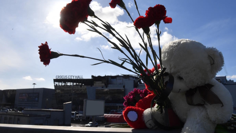 Цветы и игрушки в память о погибших при теракте на фоне сгоревшего здания концертного зала 